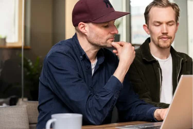 A couple of employees are focusing on a problem on a computer screen. One of the colleagues holds his chin